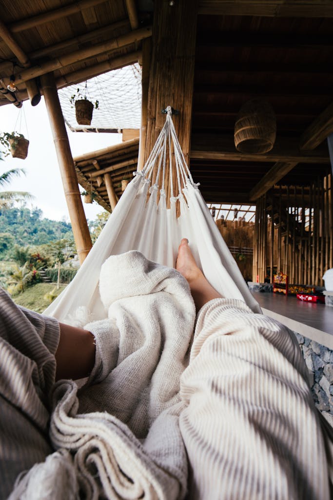 Crop barefoot traveler chilling on white cozy hammock under blanket on terrace of house in daytime